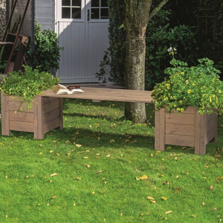 Holzbank im Garten mit zwei eingebauten Pflanzk&auml;sten voller gr&uuml;ner Pflanzen, auf einer Rasenfl&auml;che mit einem Baumstamm im Hintergrund. Vor einer wei&szlig;en T&uuml;r eines Gartenhauses oder einer Garage platziert.