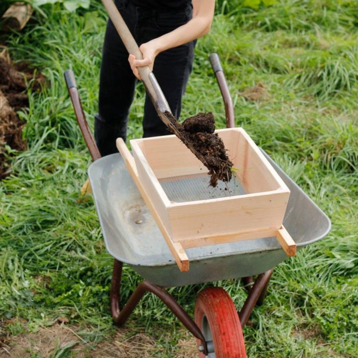 Person schaufelt Erde durch ein Holzsieb in eine Schubkarre auf einer Wiese.