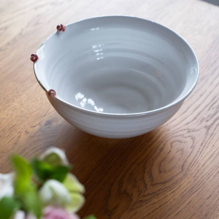 Wei&szlig;e Sch&uuml;ssel aus Keramik auf einem Holztisch, mit drei kleinen rosafarbenen Rosen am Rand verziert. Im Vordergrund unscharfe Blumen.