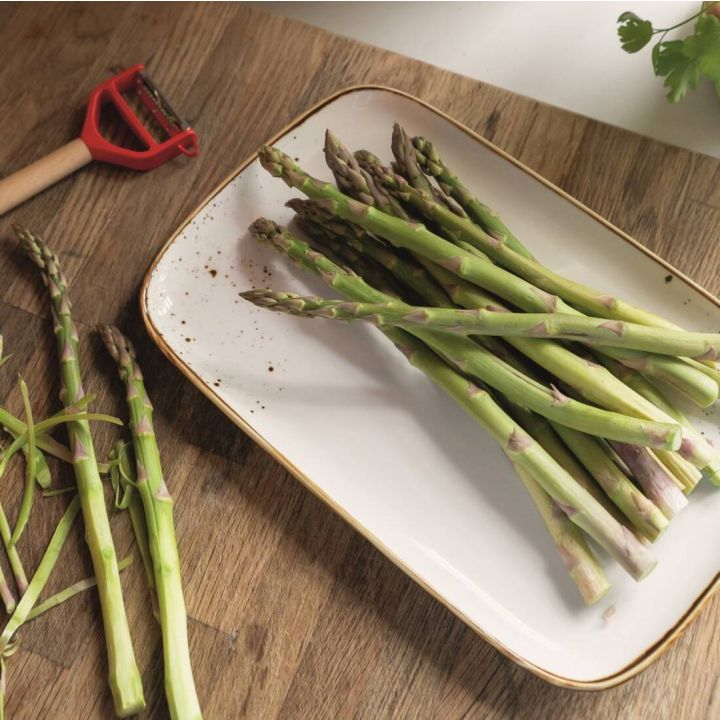 Frische grüne Spargelstangen liegen auf einem rechteckigen weißen Teller mit schmalem goldfarbenem Rand und feinen braunen Sprenkeln. Der Teller steht auf einer Holzarbeitsplatte. Links daneben befinden sich bereits geschälte Spargelstangen und ein Sparsc
