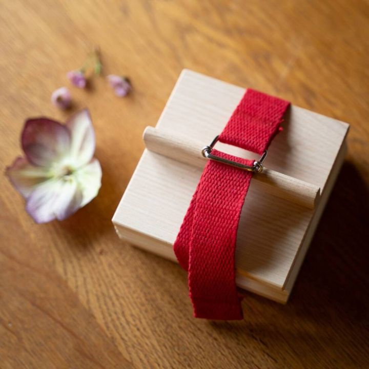 Holzbox mit rotem Textilband und Metallschnalle auf Holztisch. Neben der Box liegen eine getrocknete Blume und kleine Blütenknospen.