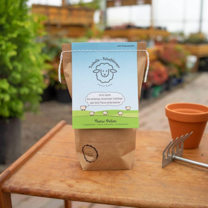 Papiertüte mit Aufschrift "Isabelle Schafdünger", gefüllt mit Naturpellets. Auf einem Holztisch stehen außerdem ein leeres Pflanztöpfchen und eine kleine Harke. Im Hintergrund Pflanzen in einem Gewächshaus.