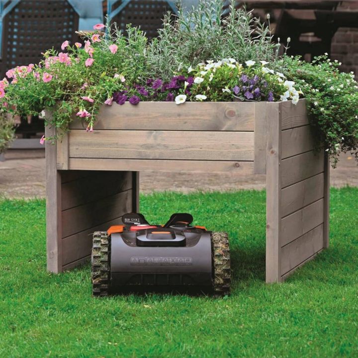 H&ouml;lzerner Pflanztisch mit bl&uuml;henden Pflanzen darauf, darunter ein Rasenm&auml;hroboter auf Rasen.