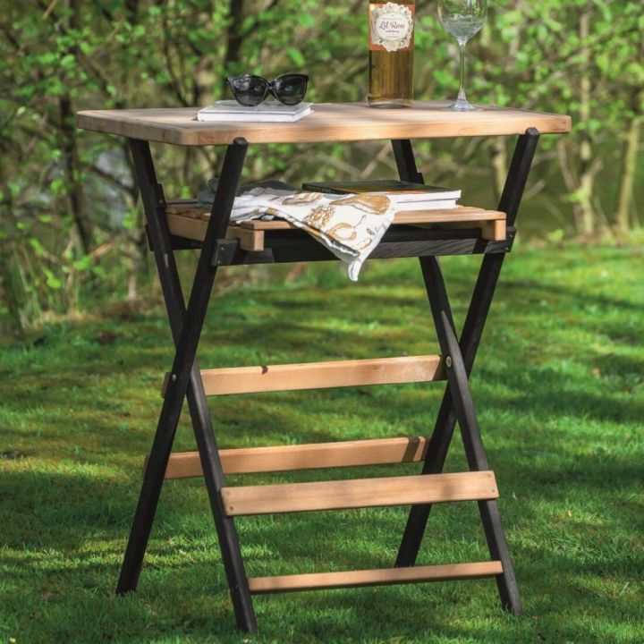 Klappbarer Beistelltisch aus Holz und Metall mit zwei Ablagefl&auml;chen im Freien; darauf stehen eine Weinflasche, ein Weinglas, eine Sonnenbrille und B&uuml;cher, ein Tuch liegt auf der unteren Ablage.