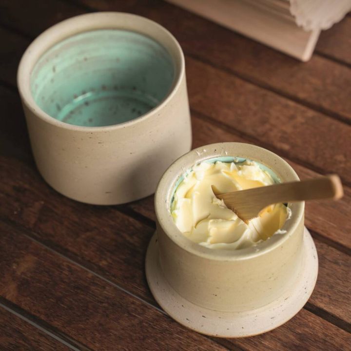 Zweiteiliges Keramik-Buttergefäß mit hellem Außendekor und türkisfarbener Glasur innen; unterer Teil enthält streichfähige Butter mit Holzspatel, oberer leerer Teil dient als Deckel.