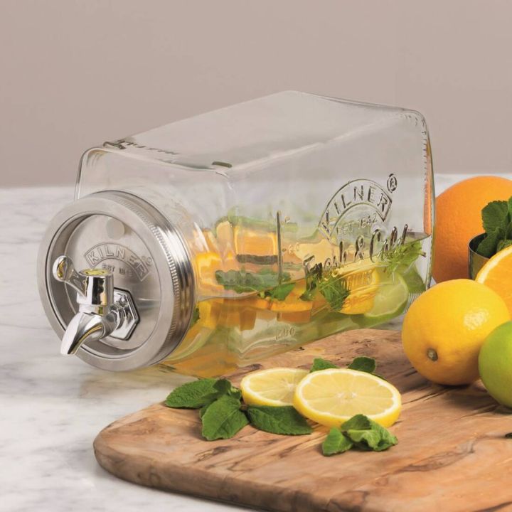Ein liegender, klarer Glasgetränkespender mit Metallhahn, gefüllt mit Wasser, Zitronen- und Limettenscheiben sowie Minzblättern, umgeben von frischen Zitronen und Limetten auf einem Holzbrett.