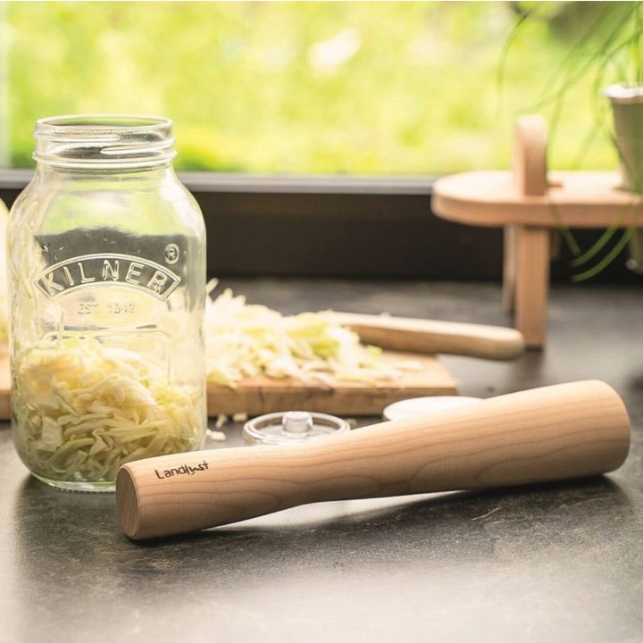 Ein Glas mit Sauerkraut der Marke &bdquo;Kilner&ldquo;, daneben ein Holzstampfer mit der Aufschrift &bdquo;Landlust&ldquo;, ein Deckel und ein Schneidebrett mit gehobeltem Wei&szlig;kohl auf einer Arbeitsplatte vor einem Fenster.