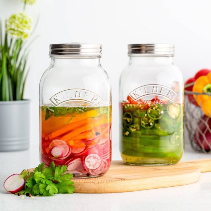 Zwei Einmachgläser von Kilner mit Gemüse, eins gefüllt mit Karotten und Radieschen in Flüssigkeit, das andere mit Paprika und Jalapeños, stehen auf einem Holzbrett. Im Hintergrund Pflanzen und ein Drahtkorb.