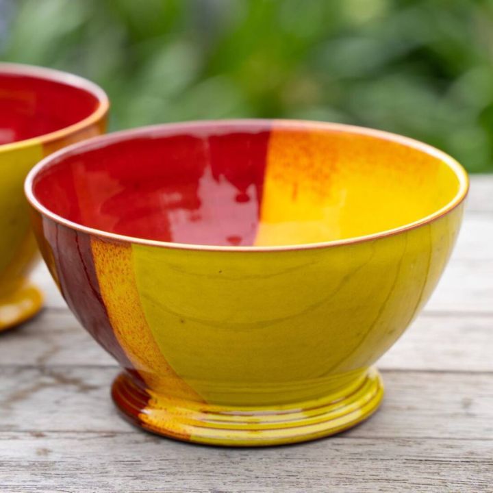 Keramikschale mit gelber und roter Glasur, stehend auf einem Holztisch. Die Innenseite ist hauptsächlich rot, die Außenseite gelb mit einem roten Bereich. Im Hintergrund verschwommene grüne Pflanzen.