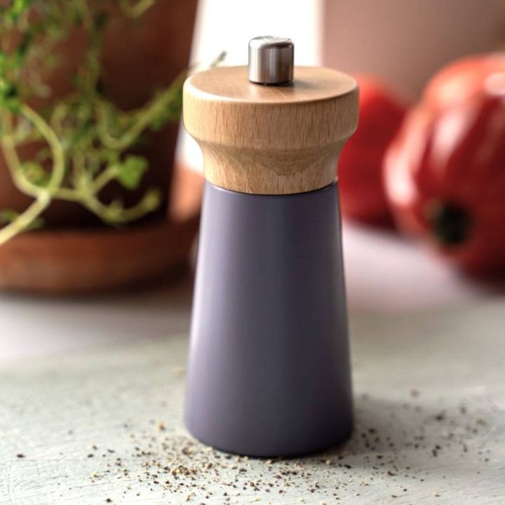 Pfeffermühle mit konischem Korpus aus mattem, dunkelgrauem Material und rundem Deckel aus hellem Holz mit Metallknopf, stehend auf heller Oberfläche mit verstreutem Pfeffer; im Hintergrund unscharfe Tomaten und Kräutertopf.