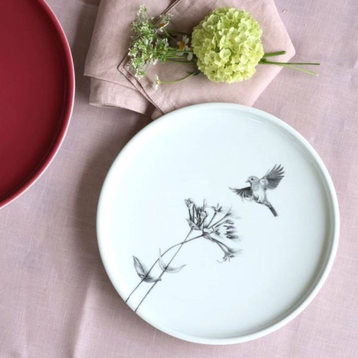 Runder, weißer Porzellanteller mit schwarz-grauer Zeichnung eines Vogels und einer Blume, daneben ein roter Teller, eine rosa Stoffserviette und ein kleiner Strauß aus grüner und weißer Blüte auf rosa Tischdecke.