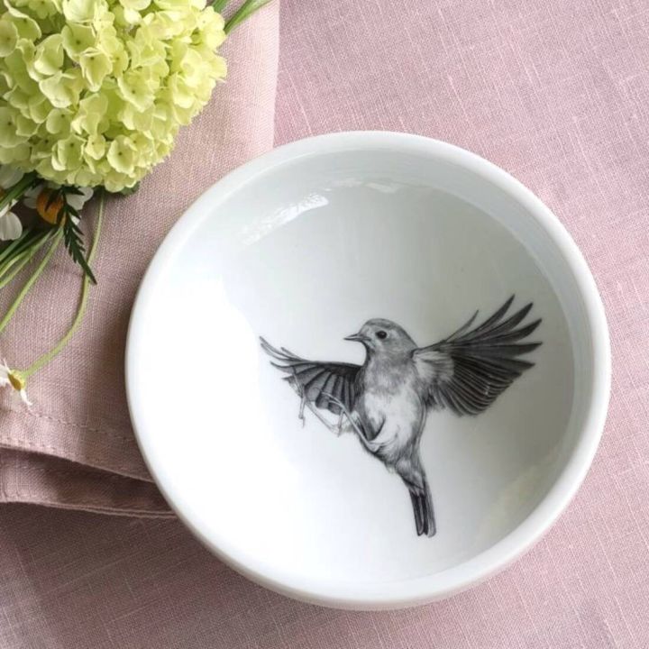 Weiße Schale mit Schwarz-Weiß-Abbildung eines fliegenden Vogels im Inneren, auf rosa Stofftuch platziert. Links oben befinden sich gelbe Blüten und grüne Blätter.