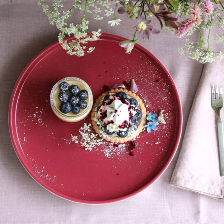 Rundes, rotes Serviertablett mit zwei Desserts auf rosa Tischdecke. Links steht ein kleines Glas mit gelber Creme, belegt mit frischen Heidelbeeren und Puderzucker. Rechts daneben eine runde Obsttarte mit Mürbeteigboden, belegt mit Heidelbeeren, Sahnehaub