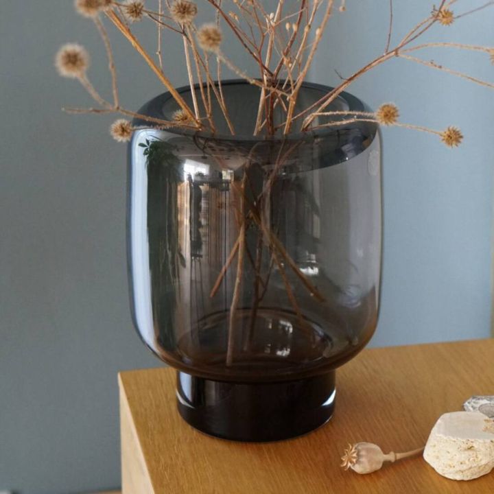 Rauchgraue Glasvase mit schwarzem Fuß steht auf einem Holztisch, gefüllt mit trockenen Zweigen, im Hintergrund blaue Wand und dekorative Objekte auf dem Tisch.