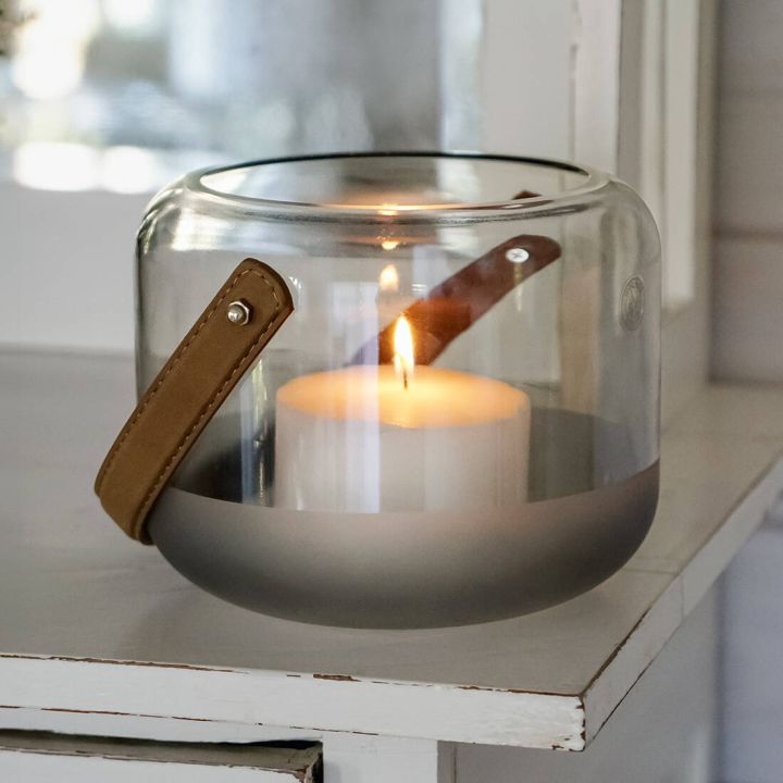 Ein Glaswindlicht mit einer brennenden wei&szlig;en Kerze darin, steht auf einem wei&szlig;en M&ouml;belst&uuml;ck. Das Glas hat eine mattierte untere H&auml;lfte und einen Lederhenkel.