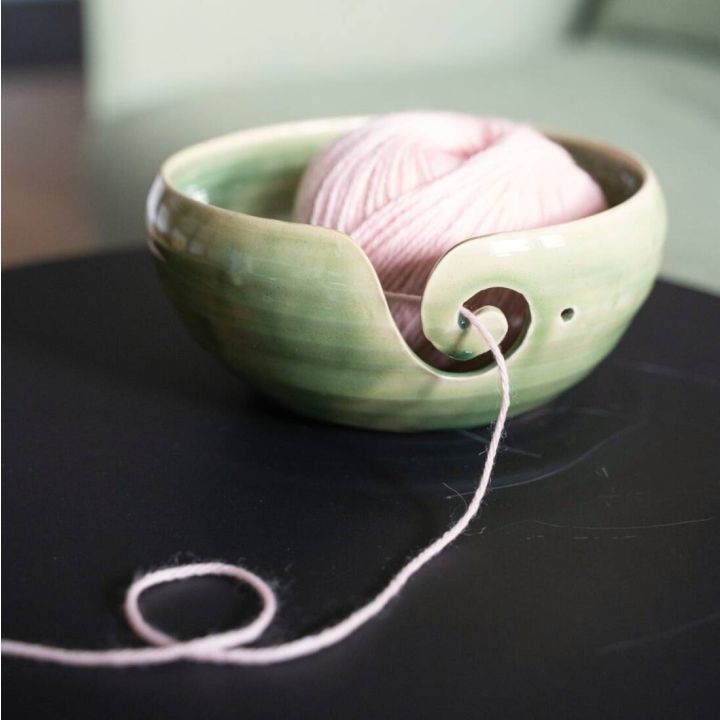 Grüne Keramikschale mit geschwungener Öffnung, hält rosa Wollknäuel. Wolle zieht durch seitlichen Schlitz, liegt auf schwarzer Oberfläche. Ideal zum ordentlichen Abwickeln beim Stricken oder Häkeln.
