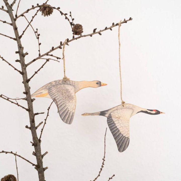Zwei fliegende Vogeldekorationen aus Papier sind an einem Ast aufgeh&auml;ngt. Die V&ouml;gel sind detailreich bemalt und h&auml;ngen an Schn&uuml;ren. Der Hintergrund ist neutral und der Ast tr&auml;gt einige kleine Zapfen.