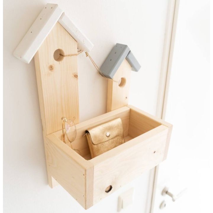 Wandorganizer aus hellem Holz mit zwei häuserförmigen Rückwänden, kleinen Dächern in Weiß und Grau, Aufhängung durch Seil, vorne ein offenes Fach mit Sonnenbrille und goldenem Etui, montiert an weißer Wand neben Tür.