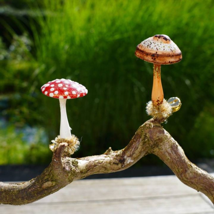 Zwei dekorative Pilzfiguren, ein Fliegenpilz mit rotem Hut und weißen Punkten sowie ein brauner Pilz, stehen auf einer gekrümmten Astnachbildung vor grünem Hintergrund.