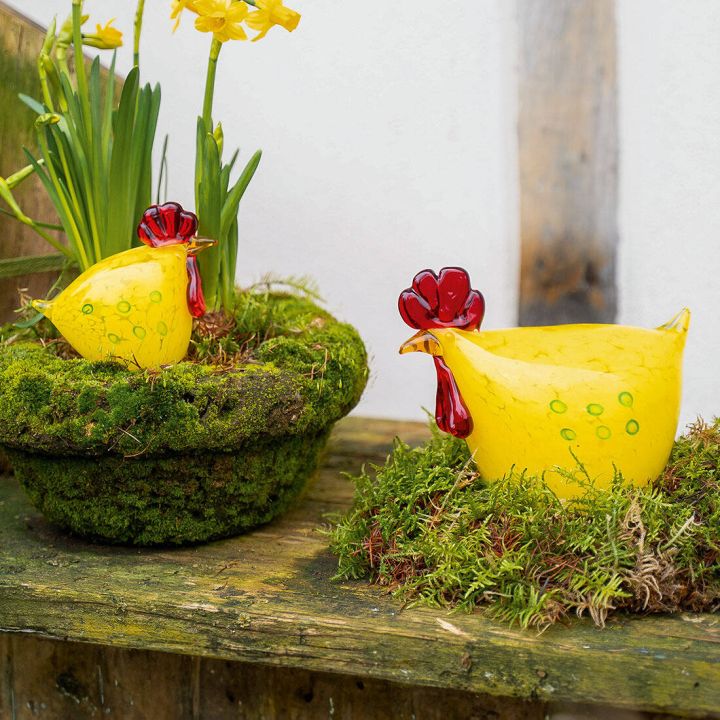 Gelbe Keramik-Hühnerfiguren mit roten Kämmen, eine sitzt in einem Mooskorb mit gelben Osterglocken, die andere steht auf Moos auf einem Holztisch.