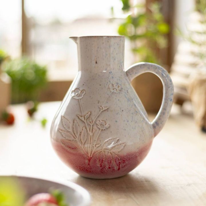 Keramikkrug mit blumenf&ouml;rmiger Pr&auml;gung und Henkel, hellgrau mit rotem Verlauf am Boden, auf einem Holztisch vor unscharfem Hintergrund mit gr&uuml;nen Pflanzen.