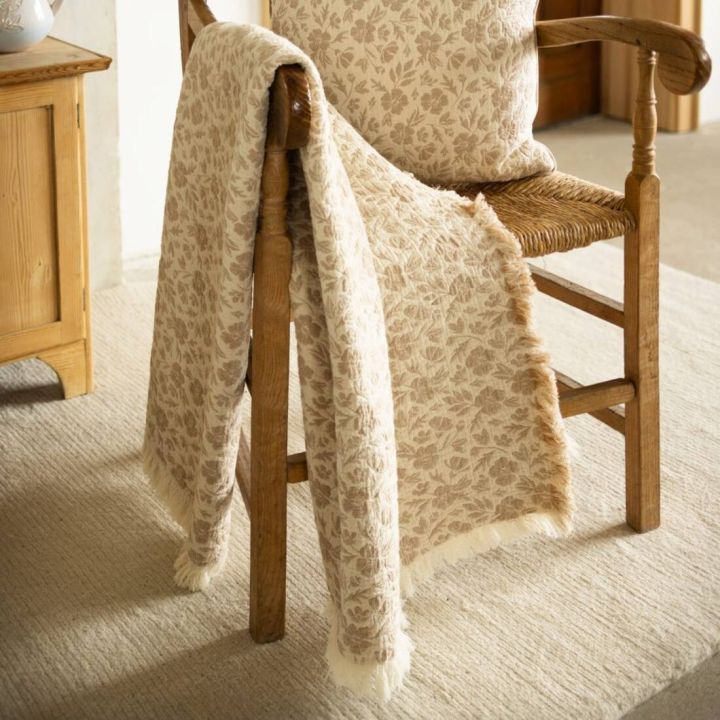 Gemusterte Decke hängt über einem Holzstuhl mit geflochtener Sitzfläche, auf dem ein passendes Kissen liegt. Beides zeigt ein florales Muster in Beige- und Brauntönen. Daneben ein Holzschrank auf einem hellen Teppich.
