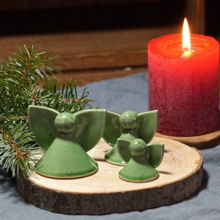 Drei gr&uuml;ne Keramikfiguren in Engelsform auf einer runden Holzscheibe, neben einem Zweig und einer brennenden roten Kerze, in gem&uuml;tlicher Umgebung arrangiert.