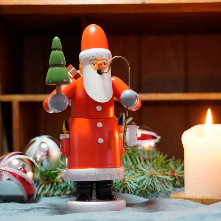 Räuchermännchen in Form eines Weihnachtsmannes mit rotem Mantel, Baum in der Hand und Pfeife. Im Hintergrund Tannenzweige und Weihnachtsschmuck. Eine brennende Kerze steht rechts auf einem Holztisch.