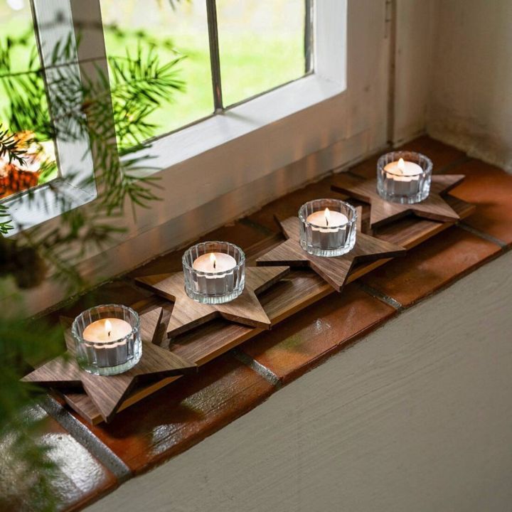 Vier brennende Teelichter in Glasbehältern stehen auf sternförmigen Holzuntersetzern auf einer Fensterbank aus roten Fliesen, dekoriert mit grünen Zweigen im Hintergrund.