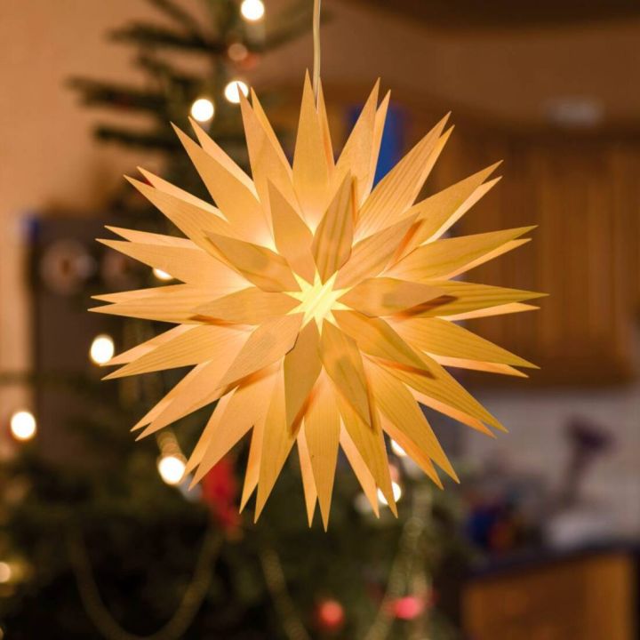 Ein leuchtender Weihnachtsstern aus Papier hängt vor einem verschwommenen Hintergrund, der einen beleuchteten Weihnachtsbaum zeigt.