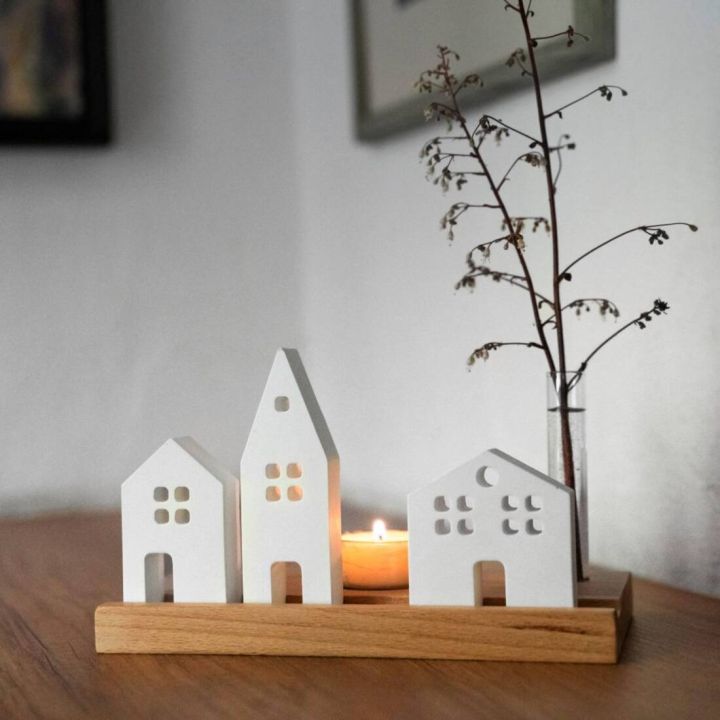 Drei weiße Keramikhäuschen mit Fensterausschnitten stehen auf einem Holzsockel, dahinter brennt ein Teelicht; rechts daneben steht ein Glas mit einem Zweig. Die Szene befindet sich auf einer Holzoberfläche vor heller Wand.