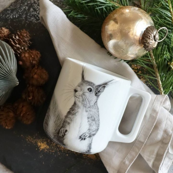 Wei&szlig;e Tasse mit Eichh&ouml;rnchen-Illustration, platziert auf beigem Stoff neben Tannenzweig und goldener Weihnachtskugel. Umgeben von Tannenzapfen und gr&uuml;nen Dekorationen auf schwarzem Untergrund.