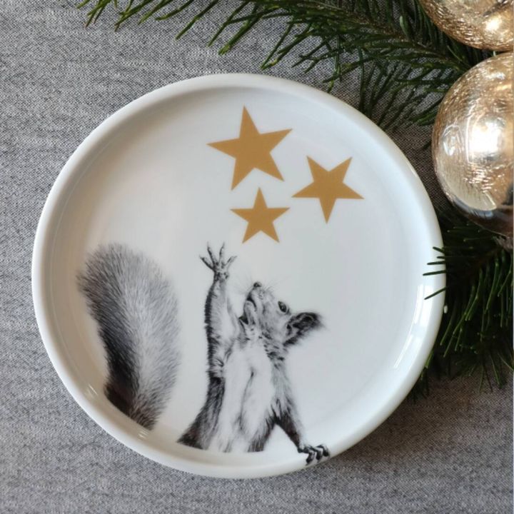 Ein runder Teller mit einem Eichhörnchenmotiv, das nach drei goldenen Sternen greift. Der Hintergrund ist grau mit Tannenzweigen und glänzenden Kugeln am Rand des Bildes.