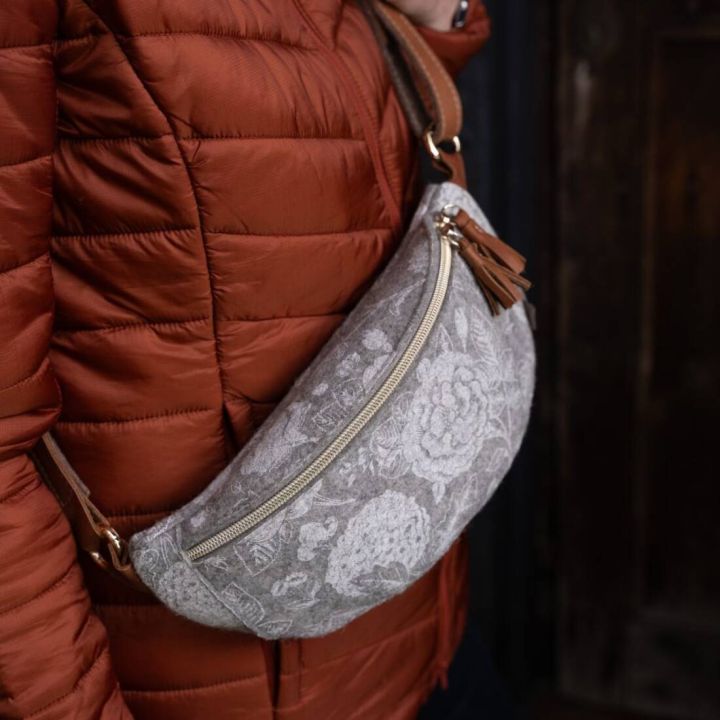 Person tr&auml;gt eine hellgraue G&uuml;rteltasche mit floralem Muster, goldenen Rei&szlig;verschluss und braunen Lederdetails &uuml;ber einer rostroten Daunenjacke.