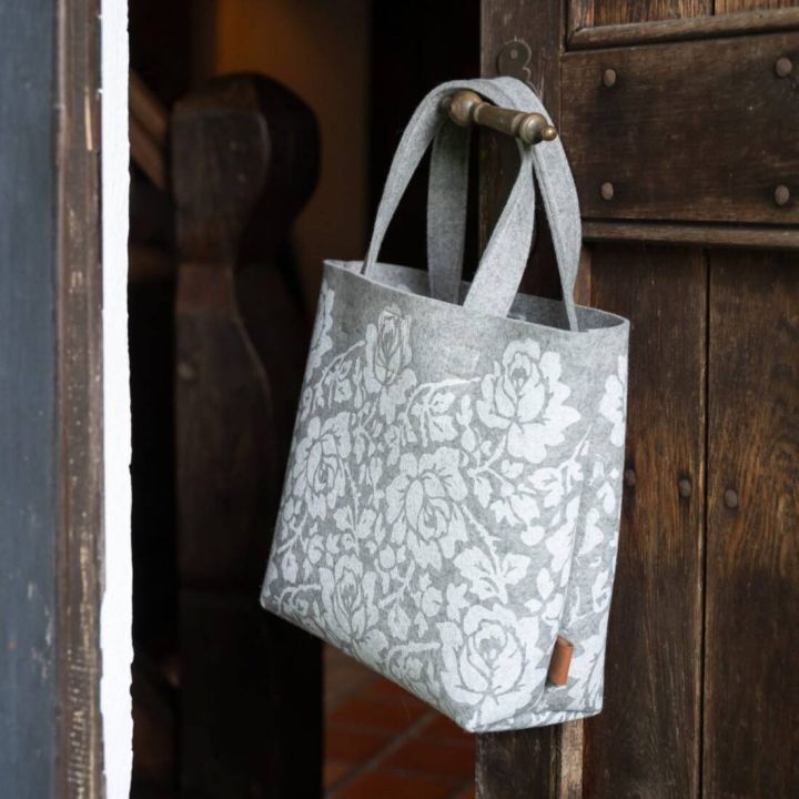 Graue Filztasche mit floralen weißen Mustern, zwei Henkeln, hängt an einer Holztürklinke neben einem Treppengeländer.