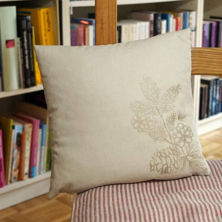 Quadratisches, beiges Kissen mit floraler Stickerei in der Ecke, auf einem Streifenstuhl vor einem Bücherregal platziert. Verschiedene Bücher in bunten Einbänden sind im Hintergrund zu sehen.