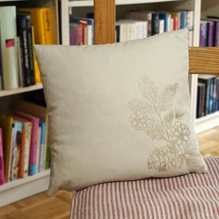 Quadratisches, beiges Kissen mit floraler Stickerei in der Ecke, auf einem Streifenstuhl vor einem B&uuml;cherregal platziert. Verschiedene B&uuml;cher in bunten Einb&auml;nden sind im Hintergrund zu sehen.