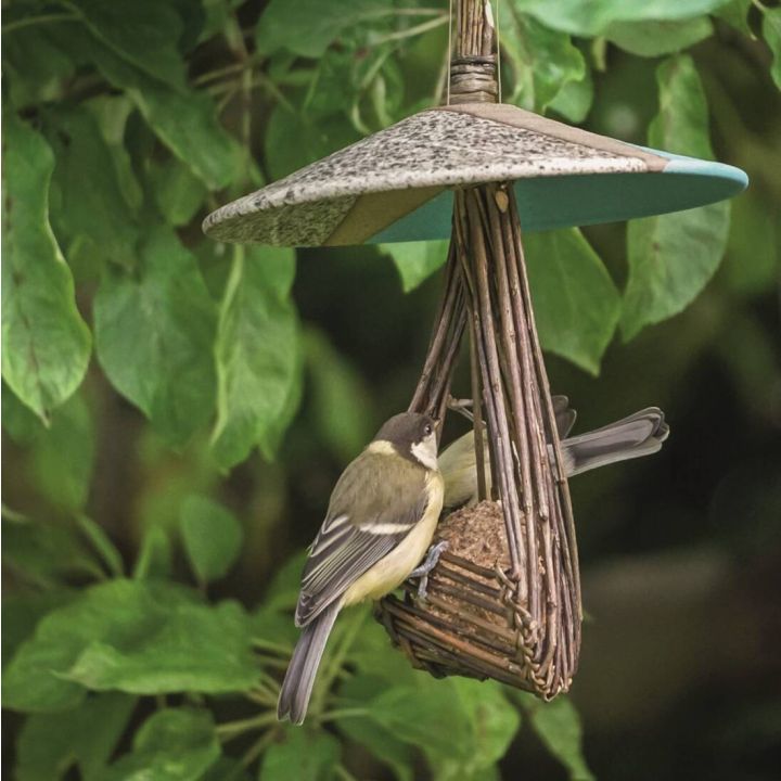 Vogelfutterhaus aus Holz mit zwei V&ouml;geln, die an einem Futterball picken. Das Haus hat ein rundes Dach und h&auml;ngt vor gr&uuml;nen Bl&auml;ttern.
