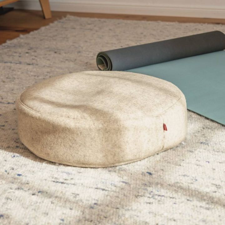 Meditationskissen aus beigem Material auf einem Teppich, neben einer aufgerollten Yoga-Matte. Das Kissen hat eine runde Form und eine glatte Oberfläche.