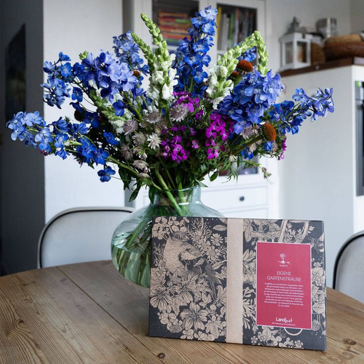 Gro&szlig;er Blumenstrau&szlig; in Vase mit blauen, lila, wei&szlig;en Bl&uuml;ten. Daneben dekorative Produktverpackung mit floralem Muster und rotem Etikett auf einem Holztisch. Helle, moderne Inneneinrichtung im Hintergrund.