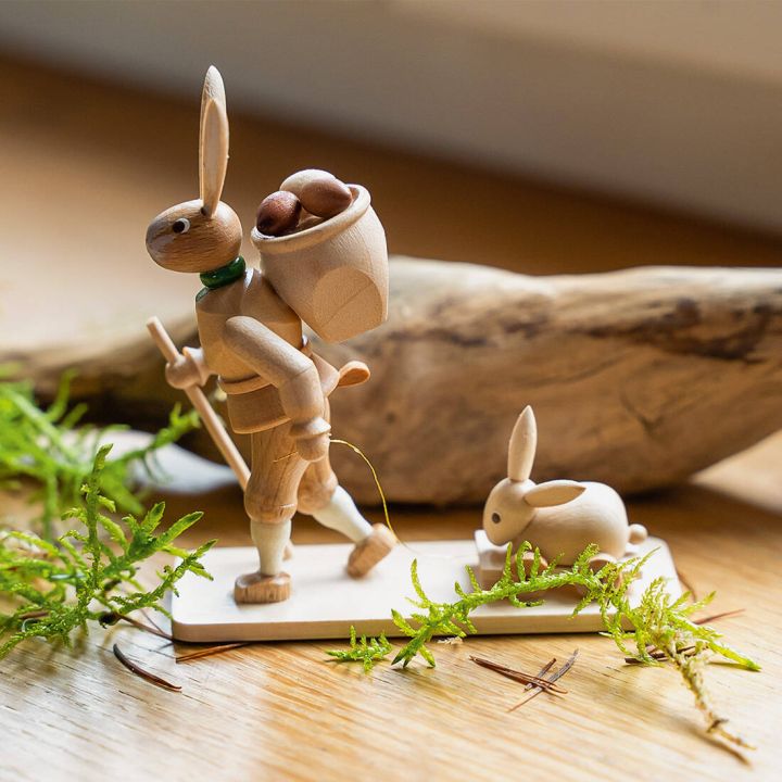 Holzfigur eines gehenden Hasen mit Rucksack voller Eier und Wanderstock, führt auf einer rechteckigen Bodenplatte einen kleinen Hasen auf einem Schlitten; Dekoration mit Moos und Holzstück im Hintergrund.