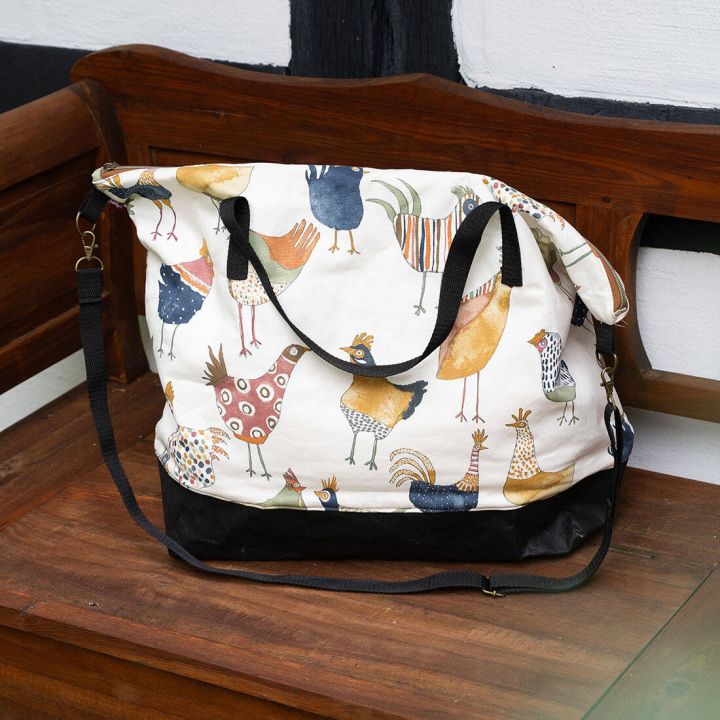 Wei&szlig;e Stofftasche mit bunten, stilisierten H&uuml;hnern; schwarze Tr&auml;ger und Boden; sitzt auf einer Holzbank.