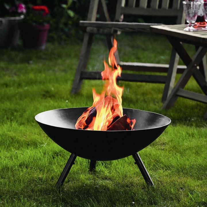Schwarze Feuerschale auf Rasen stehend, mit brennendem Holz und Flammen, umgeben von Gartenmöbeln im Hintergrund.