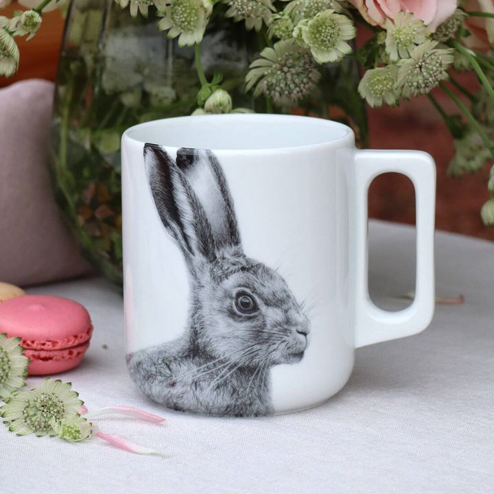 Wei&szlig;e Tasse mit einem detaillierten Schwarz-Wei&szlig;-Zeichnung eines Hasenkopfes. Die Tasse steht auf einem Tisch neben rosa Macarons und einem Blumenarrangement mit gr&uuml;nen und rosa Bl&uuml;ten.