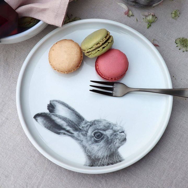 Ein Teller mit einem Hasenmotiv, darauf drei Macarons in beige, gr&uuml;n und rosa, liegt auf einer grauen Tischdecke. Daneben liegt eine silberne Gabel.