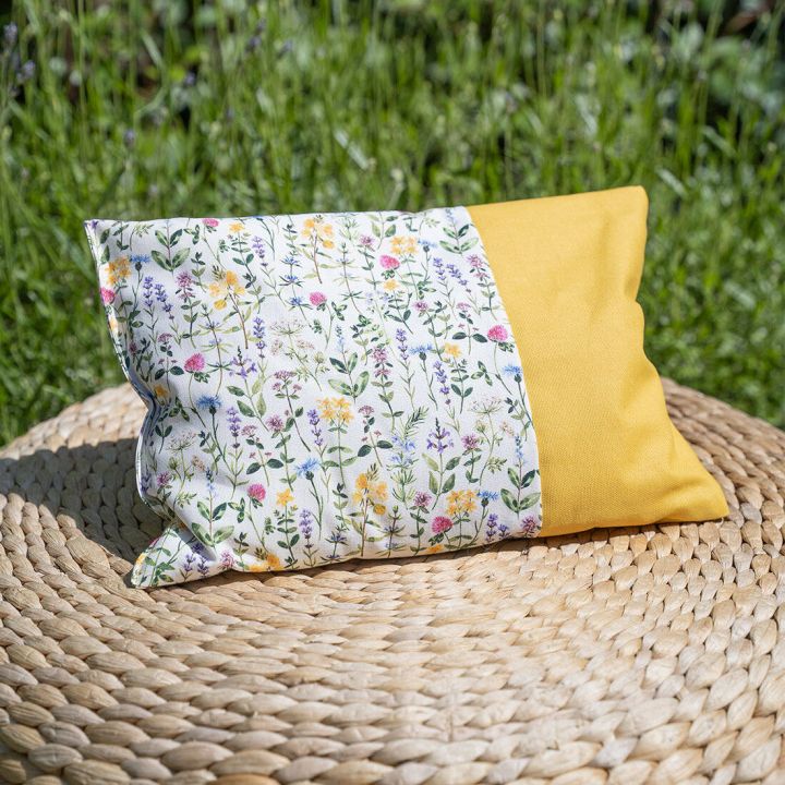 Buntes Kissen mit gelber Seite und floraler Musterung, liegend auf geflochtener runder Unterlage im gr&uuml;nen Garten.