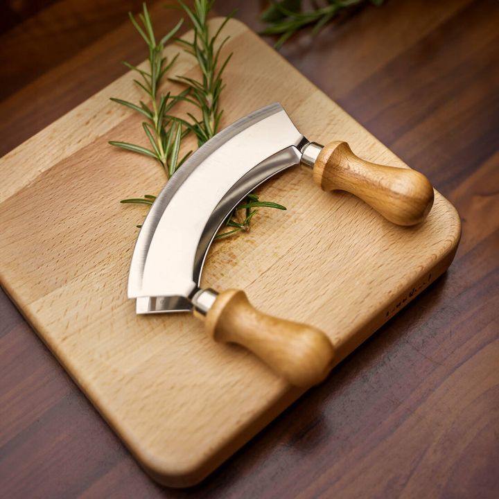 Halbrundes Wiegemesser mit zwei Holzgriffen liegt auf einem rechteckigen Holzschneidebrett. Unter dem Messer befinden sich einige Zweige frischer Rosmarin. Das Schneidebrett hat eine Mulde in der Mitte und liegt auf einer dunklen Holzoberfl&auml;che.