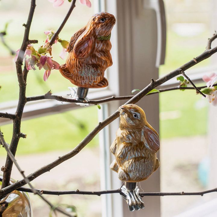 Zwei dekorative Hasenfiguren aus Glas sind auf bl&uuml;henden &Auml;sten in einem Raum mit gro&szlig;en Fenstern befestigt. Die Hasen sind detailliert gestaltet und funkeln leicht im Licht.
