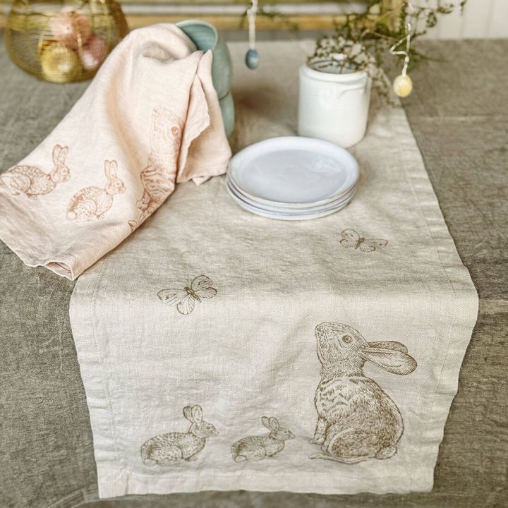 Tischl&auml;ufer und Geschirrtuch aus Leinen mit aufgedruckten Hasen und Schmetterlingen, dekorativ auf einem gedeckten Tisch mit Tellern, Tassen und Vase arrangiert. Im Hintergrund Osterdekoration mit Eiern in einem Drahtkorb.