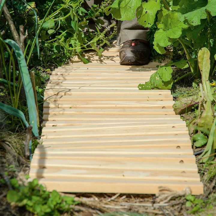 Holzsteg aus schmalen Brettern führt durch grünen Garten, Schuhe einer Person am oberen Rand sichtbar, Pflanzen überwuchern seitlich den Steg.